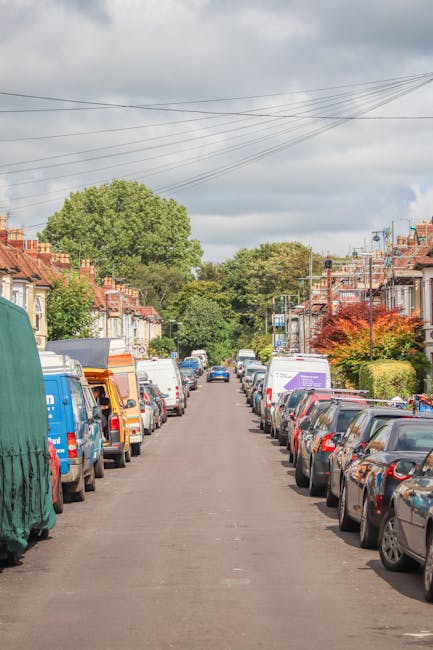 A residential street during daytime with parked cars lining both sides of the narrow asphalt road. The vehicles include various hatchbacks, sedans, and vans, some of which belong to local businesses with visible branding. Behind the parked cars, there are terraced houses with brick facades, small front gardens, and pitched roofs, some featuring chimneys. A few trees and shrubs add greenery along the pavement, and the sky above is partly cloudy with patches of sunlight. Overhead, power lines run parallel to the street, supported by wooden utility poles. The scene depicts a typical quiet neighbourhood environment suitable for home relocation or furniture transport activities, with no moving activity visible but indicative of an area where professional removals services may operate, such as those offered by Man with Van South Woodford.