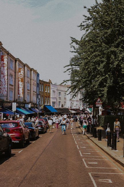 A busy street scene on South Woodford High Road showing parked cars lining both sides of a one-way road with white parking box markings. On the right side, a row of multicoloured terraced houses and shops with blue and white awnings can be seen, while pedestrians are walking along the pavement, some carrying boxes or bags, suggesting a relocation or moving activity. A large leafy tree extends over the street, partially shading the area. In the background, a van from Man with Van South Woodford is parked near a loading zone, with furniture, cardboard boxes, and packing materials visible inside and around the vehicle, indicating the process of home relocation and furniture transport. The scene is well-lit, capturing the typical urban environment used for moving and removals services in South Woodford.