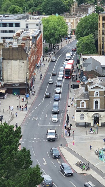 An aerial view of South Woodford High Road showing parked cars along the curb and moving vehicles on the road, with pedestrians walking on the pavements and some gathering near small shops and commercial buildings. The street is lined with leafy green trees providing partial shade, and residential houses with pitched roofs are visible further along the street. The scene appears to be during daytime with clear weather. This setting exemplifies the typical environment where Man with Van South Woodford might perform house removals or furniture transport, highlighting the importance of suitable parking spots for an efficient loading process during home relocation or packing and moving services. The image captures the urban street environment where strategic parking enables smooth logistics for residential removals in South Woodford.