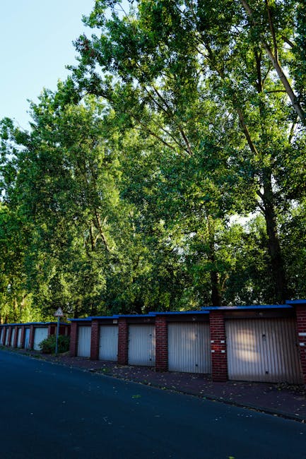 A busy street scene on South Woodford High Road showing parked cars lining both sides of a one-way road with white parking box markings. On the right side, a row of multicoloured terraced houses and shops with blue and white awnings can be seen, while pedestrians are walking along the pavement, some carrying boxes or bags, suggesting a relocation or moving activity. A large leafy tree extends over the street, partially shading the area. In the background, a van from Man with Van South Woodford is parked near a loading zone, with furniture, cardboard boxes, and packing materials visible inside and around the vehicle, indicating the process of home relocation and furniture transport. The scene is well-lit, capturing the typical urban environment used for moving and removals services in South Woodford.