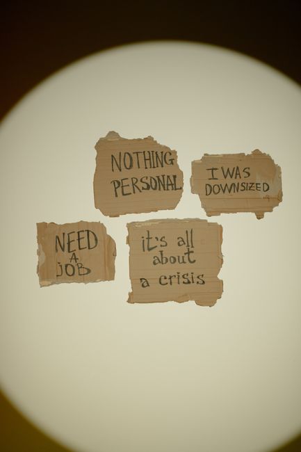 A close-up view of four torn pieces of cardboard taped to a light-colored wall, each displaying handwritten messages in black ink. The messages read 'NOTHING PERSONAL,' 'I WAS DOWNSIZED,' 'NEED A JOB,' and 'it's all about a crisis.' The torn edges of the cardboard suggest they may be part of a protest or personal expression. The lighting creates a circular spotlight effect around the messages, emphasizing their prominence. This image is relevant to themes of personal and professional upheaval, which can be connected to home relocations and moving services managed by companies like Man with Van South Woodford, particularly when individuals are facing significant life changes such as downsizing or job loss before a move.