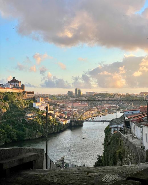 A scenic view of a river flowing through an urban area during sunset, with a historic building on a hilltop to the left and modern high-rise buildings in the distance. In the foreground, there are rooftops and a narrow street, with some greenery on the slopes beside the river. The sky is partly cloudy with a mix of light and darker clouds, casting a warm glow over the scene. This image captures a moment during a home relocation or house move, highlighting urban landscape and architectural elements typical in South Woodford, relevant to the topic of house removals and moving logistics facilitated by Man with Van South Woodford.