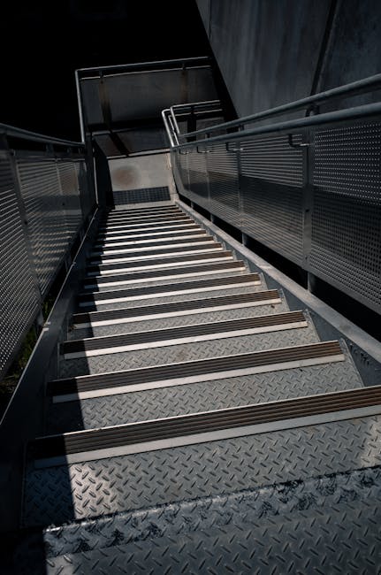 A metal staircase with grated steps ascends from an outdoor area to an enclosed building entrance, featuring dark metal railings and transparent panels on either side. The stairs are illuminated by natural daylight, casting shadows on the treads. The surrounding environment suggests an urban setting, with the staircase providing access to a higher level, likely for residential or commercial purposes. This type of access may present challenges during home removal or furniture transport if narrow or difficult to navigate. At the top of the stairs, a doorway or entrance may be visible, indicating entry into a property or building. Man with Van South Woodford, a professional removal company, specializes in handling logistics for home relocations, including navigating narrow stairs and access issues like this. Their services typically involve careful packing and transportation of furniture and boxes from such challenging entry points to their vehicle for efficient home removal or moving operations.
