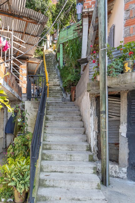 A narrow residential alleyway on George Lane in South Woodford, showing paved concrete steps leading uphill between tall brick and white-painted houses. On the right side, a brick wall with a street sign reading 'FALLERS LANE' is visible. The left side features a white building with small, rectangular windows and a black external drainpipe running vertically down the wall. The alley is illuminated by natural daylight, with a streetlamp positioned on the upper part near the steps and some greenery visible at the top of the incline. This setting illustrates a typical challenging access route for home relocations involving narrow stairs and restricted space, where professional removals, such as those by Man with Van South Woodford, would carefully navigate furniture and boxes along such pathways as part of their furniture transport and packing and moving services.
