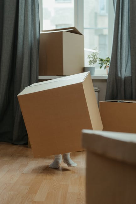 Inside a residential room with natural daylight coming through a large window adorned with dark curtains, several large cardboard boxes are arranged on a wooden floor, some placed on top of a radiator