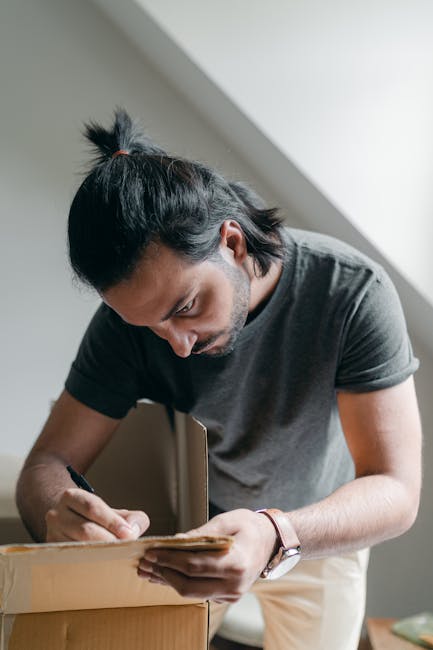 A man with dark hair tied in a small ponytail, wearing a dark grey t-shirt, is inside a residential property, packing belongings into a cardboard box. He is focused on sealing or inspecting the box, which is partially filled with paper and packing materials. The background shows a plain light-coloured wall and natural lighting coming from an unseen window, indicating an indoor environment during a home relocation process. This scene depicts the packing stage of furniture transport and house removals, with attention to careful handling and organisation, consistent with services offered by Man with Van South Woodford.