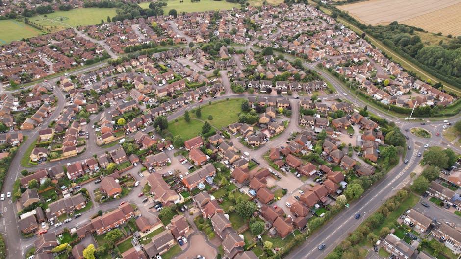 An aerial view of a residential neighbourhood within Snaresbrook Estate in South Woodford, showing numerous detached and semi-detached houses with pitched roofs, surrounding a central green park area with a few trees. The streets are lined with parked cars and are winding through the housing development, with a main road visible along the estate's perimeter, featuring moving vehicles. To the right, there are agricultural fields and patches of green space, indicating the area's suburban setting. The image captures the layout of the estate during daytime with natural lighting, illustrating the typical environment where house removals and furniture transport services by Man with Van South Woodford are often required, especially during home relocations involving packing, loading, and moving furniture and boxes from houses to moving vans.