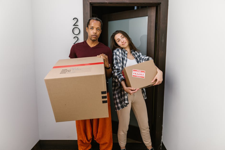 A man with dark hair tied in a small ponytail, wearing a dark grey t-shirt, is inside a residential property, packing belongings into a cardboard box. He is focused on sealing or inspecting the box, which is partially filled with paper and packing materials. The background shows a plain light-coloured wall and natural lighting coming from an unseen window, indicating an indoor environment during a home relocation process. This scene depicts the packing stage of furniture transport and house removals, with attention to careful handling and organisation, consistent with services offered by Man with Van South Woodford.