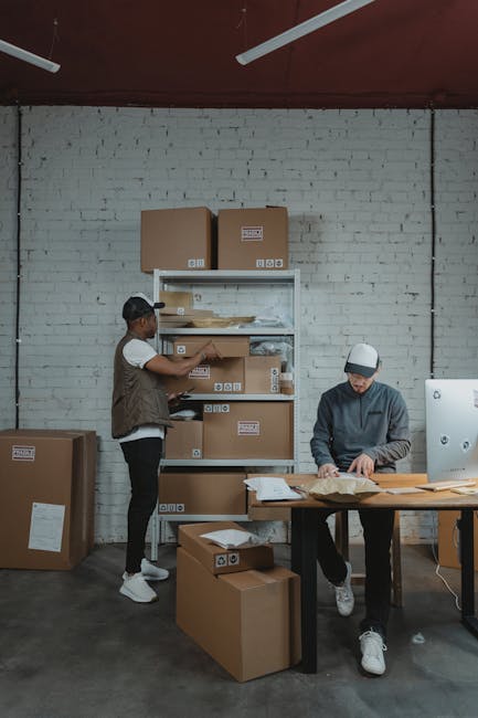 Inside a storage room with white brick walls and a concrete floor, two men are engaged in packing and organizing boxes for house removals. One man is standing on the left, wearing a dark vest and cap, handling a cardboard box on a metal shelving unit that holds additional boxes and packing materials. The other man is seated on the right, wearing a hoodie and white cap, working on a desk with papers, packing supplies, and a computer. Surrounding them are several sealed cardboard boxes of various sizes, some stacked on the floor, indicating the process of packing items for a home relocation. The scene is lit by natural and artificial light, capturing the careful handling of packing materials and the structured environment typical of professional removals and storage services, as provided by Man with Van South Woodford.