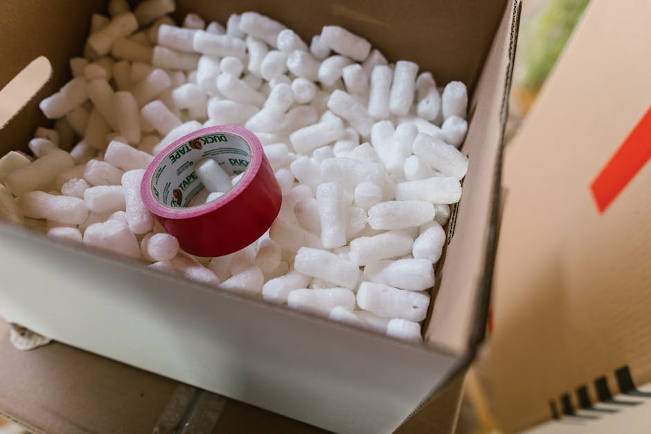Close-up view of a cardboard moving box labeled 'WINTER CLOTHS' in bold black letters, with clear plastic packing tape sealing the top. A person's hand is seen pressing down slightly on the box while another person, wearing blue jeans and a beige sweatshirt, is reaching over the box, possibly for additional tape or to stabilize it. The setting appears to be inside a property or a storage area, with additional boxes visible in the background, also labeled for packing categories. The boxes are arranged on a surface, likely for the packing process during a home relocation or moving service. This scene captures the detailed elements involved in packing belongings securely for transport, a key part of professional removals services offered by companies such as Man with Van South Woodford. The environment suggests an organized packing session, with various tools and materials, including scissors, visible nearby to facilitate efficient moving and packing procedures.