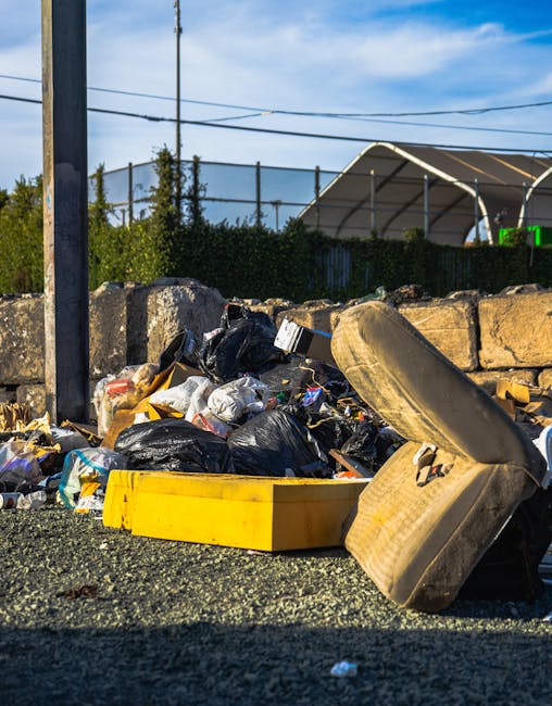 A large pile of household waste and discarded items positioned outdoors on a gravel surface next to a low stone wall, with black garbage bags, plastic packaging, and various rubbish scattered around. An old, worn-out mattress leans against the stone wall on the right side, partially blocking some of the waste. A yellow plastic container is visible on the ground among the rubbish. In the background, there are metal power lines, a tall utility pole, and a green hedge behind the stone wall, with a partly cloudy blue sky above. The setting appears to be an outdoor waste collection or disposal area, relevant to house removals and packing services offered by Man with Van South Woodford, who handle disposal and logistics as part of their overall house removals and furniture transport offerings.