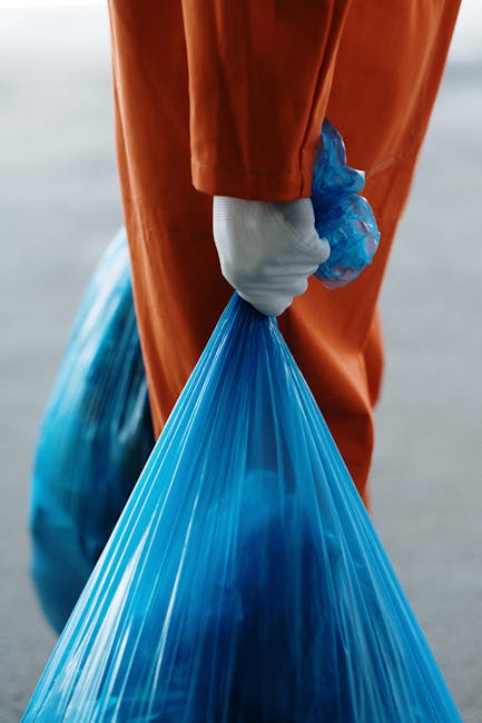 A person wearing an orange warehouse uniform and white gloves is holding a large blue plastic bag filled with waste material, standing on a paved surface. In the background, there are more similar blue bags, indicating a collection of waste for removal. The scene appears to be part of a home or commercial waste disposal process, with the individual ready to transport or load the bags. This image illustrates aspects of waste collection associated with house removals or clutter clearance services, such as packing or disposing of bulky waste during a home relocation. The lighting is natural, highlighting the plastic bags and the worker’s attire, emphasizing efficient and organized waste handling in a professional removal setting, possibly managed by companies like Man with Van South Woodford.