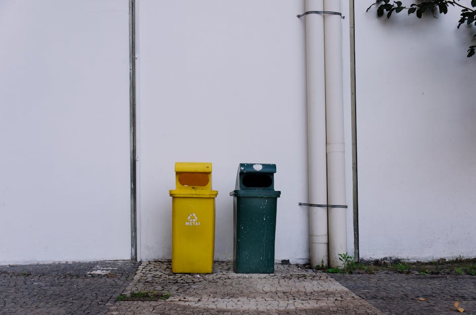A large pile of household waste and discarded items positioned outdoors on a gravel surface next to a low stone wall, with black garbage bags, plastic packaging, and various rubbish scattered around. An old, worn-out mattress leans against the stone wall on the right side, partially blocking some of the waste. A yellow plastic container is visible on the ground among the rubbish. In the background, there are metal power lines, a tall utility pole, and a green hedge behind the stone wall, with a partly cloudy blue sky above. The setting appears to be an outdoor waste collection or disposal area, relevant to house removals and packing services offered by Man with Van South Woodford, who handle disposal and logistics as part of their overall house removals and furniture transport offerings.