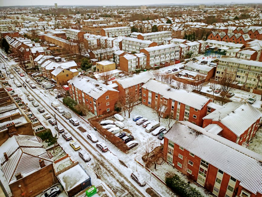 An aerial view of a residential neighbourhood in South Woodford during winter, showing snow-covered rooftops, streets, and parked cars. Brick apartment buildings and houses with snow on their roofs are visible, along with a lined street where several cars are parked along the curb. The street appears to be part of a home relocation or moving scene, with some vehicles and activity possibly associated with furniture transport or packing and moving processes. The environment is well-lit with natural daylight, highlighting the snow and the uniformity of the urban layout, emphasizing the typical setting for local house removals services offered by Man with Van South Woodford.