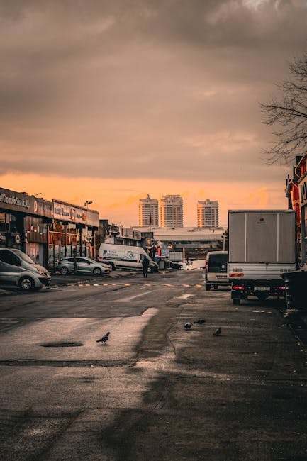 The image depicts a parking area outside a commercial building during sunset, with a mostly cloudy sky casting an orange hue over the scene. In the foreground, a white van is being loaded with cardboard moving boxes of various sizes, some wrapped in plastic or fabric for protection. A person is visible, lifting and carrying a box towards the vehicle, while a few pigeons are walking nearby on the wet asphalt surface. Behind the loading activity, additional vehicles including cars and a small truck are parked along the pavement. To the left, the shopfronts display signage, and on the right side, there is a building facade. In the background, three high-rise residential buildings stand under the sunset sky, indicating an urban area. This scene is representative of a home relocation process, involving the packing, handling, and transportation of household items by [COMPANY_NAME], a professional removals service specializing in house moves and furniture transport in the E18 area.